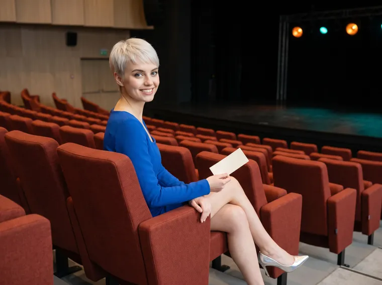 Generated image: the site character sits in an aisle seat of an empty concert hall with rust-red seats before the doors open, holding a plain paper ticket.